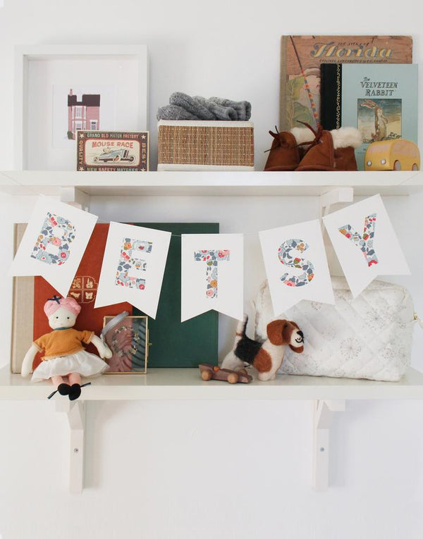 Bunting personalised with child's name in Liberty Betsy fabric hanging on nursery shelf.